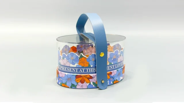 We offer a custom cylindrical packaging solution featuring a clear body with a blue handle and adorned with an elegant floral pattern in orange, blue, and purple hues. This portable container is crafted for versatility and is ideal for various gift-giving occasions, providing both aesthetic appeal and practicality.