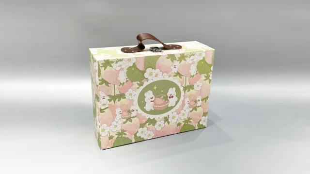 Our product is a custom box packaging featuring a floral and bunny design. It includes a durable brown leather handle and is secured with a sleek white clasp. The front of the box showcases a circular logo emblem, adding a touch of elegance. This portable gift box is presented against a smooth, gray backdrop to highlight its quality craftsmanship and aesthetic appeal.