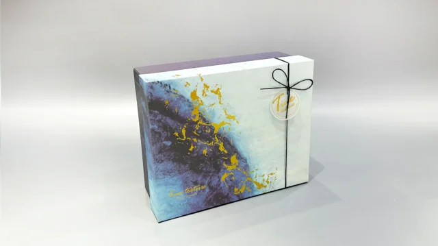 This custom ribbon box showcases a striking blue and gold abstract pattern. Designed with a lift-off lid, it is elegantly finished with a black cord and includes a round tag for added personalization.