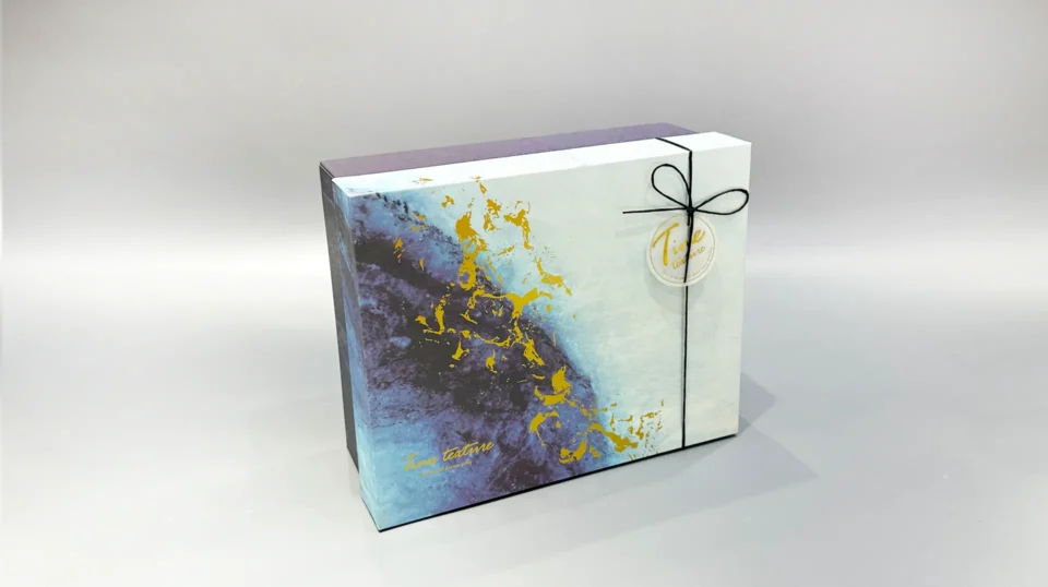 This custom ribbon box showcases a striking blue and gold abstract pattern. Designed with a lift-off lid, it is elegantly finished with a black cord and includes a round tag for added personalization.