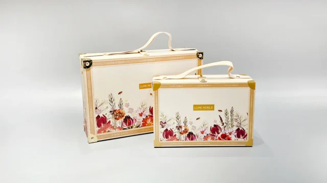 We are a manufacturer of custom box packaging. This image represents our product. Please revise the description to: "Two cream-colored suitcase-style boxes with intricate floral patterns, featuring elegantly embossed decorative elements. These portable, flip-top boxes are designed to be both stylish and functional, making them ideal for secure travel or home storage solutions.