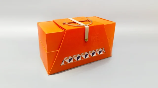 This image showcases our custom-designed packaging: a high-end, geometric orange box crafted from cardboard. It features a convenient handle and a sophisticated triangular pattern on one side, exuding the elegance of a portable gift box. The backdrop is minimalist, emphasizing the luxurious and unique design of our product.