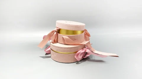 Our product image showcases three custom round gift boxes in a soft pink hue, each embellished with elegant gold bands and topped with neatly tied pink ribbons. These boxes are artfully arranged in a stacked formation against a neutral background, emphasizing the sophisticated design and luxurious appeal of our packaging solutions.