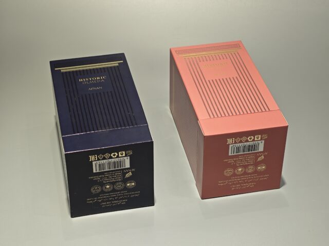 Two rectangular perfume boxes are displayed side by side on a light background. The left box features a dark color scheme with elegant UV printing that highlights its sophisticated design. The right box has a pink hue with gold detailing, both exemplifying exquisite handmade craftsmanship in their construction and finish.