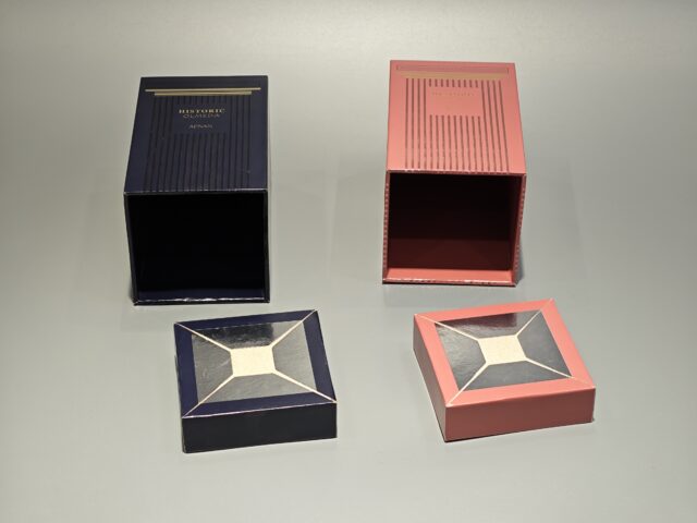 Two rectangular perfume boxes are displayed side by side on a light background. The left box features a dark color scheme with elegant UV printing that highlights its sophisticated design. The right box has a pink hue with gold detailing, both exemplifying exquisite handmade craftsmanship in their construction and finish.
