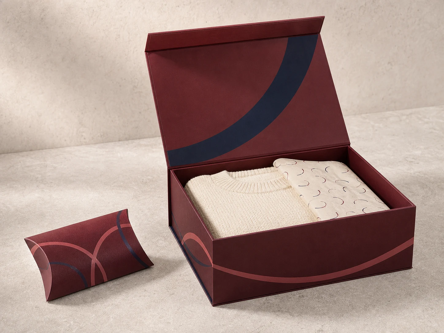 Open oxblood magnetic garment box with folded ivory knitwear, patterned tissue paper, and a matching pillow box.