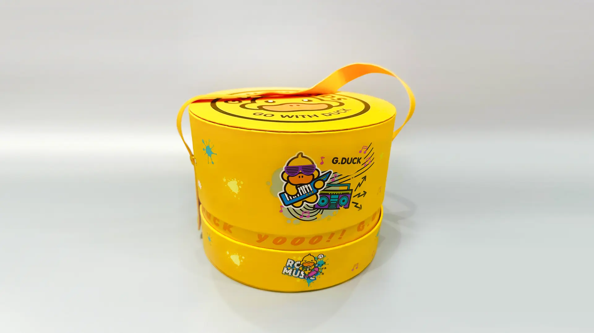 A yellow, round, two-layered gift box featuring a playful cartoon duck and boombox design is equipped with an orange carrying handle. This portable packaging solution is ideal for transporting lunches or small gifts.