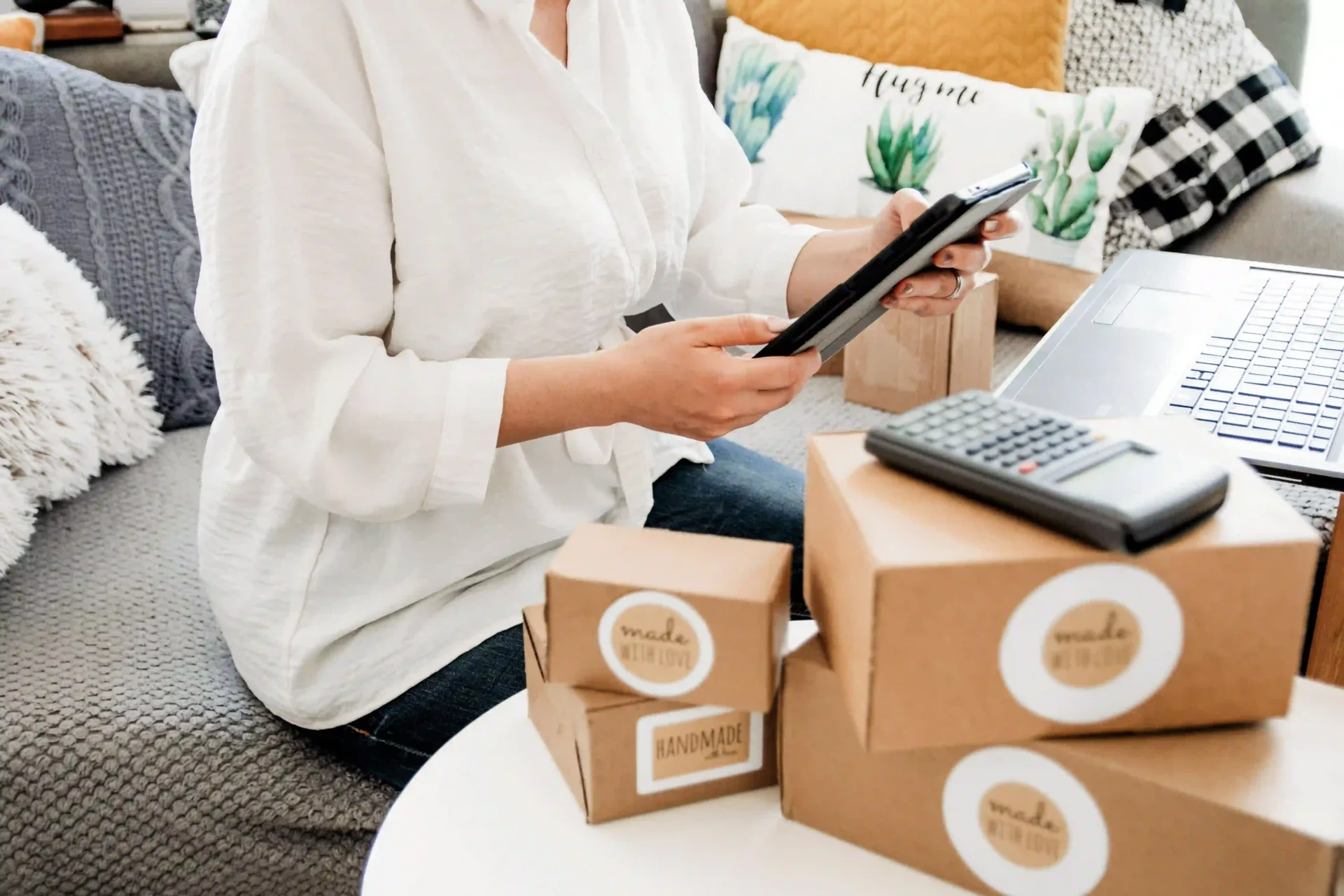 A person is holding a tablet next to several cardboard boxes with visible labels, with a calculator on the table, suggesting a small business or home office setting. The focus is on the personalized packaging solutions, emphasizing the variety of sizes and shapes of the boxes without specifying any text on them.
