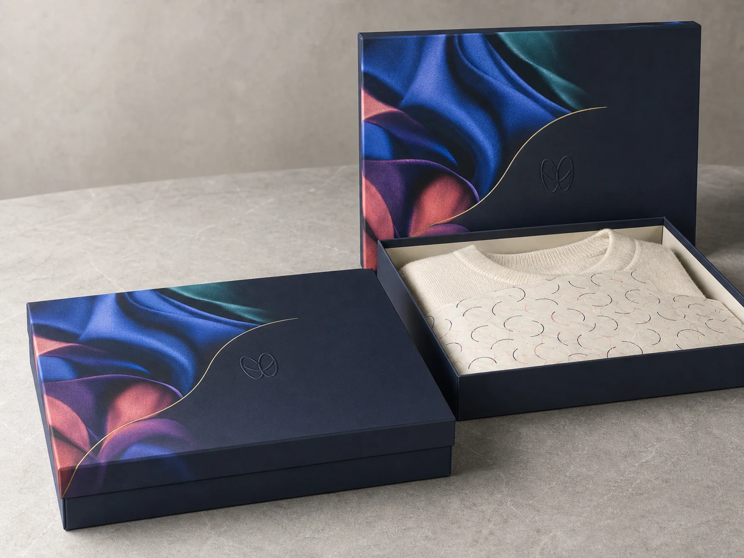 Navy rigid garment boxes, one open and one closed, with colorful fabric print, gold foil accents, knitwear, and tissue paper.