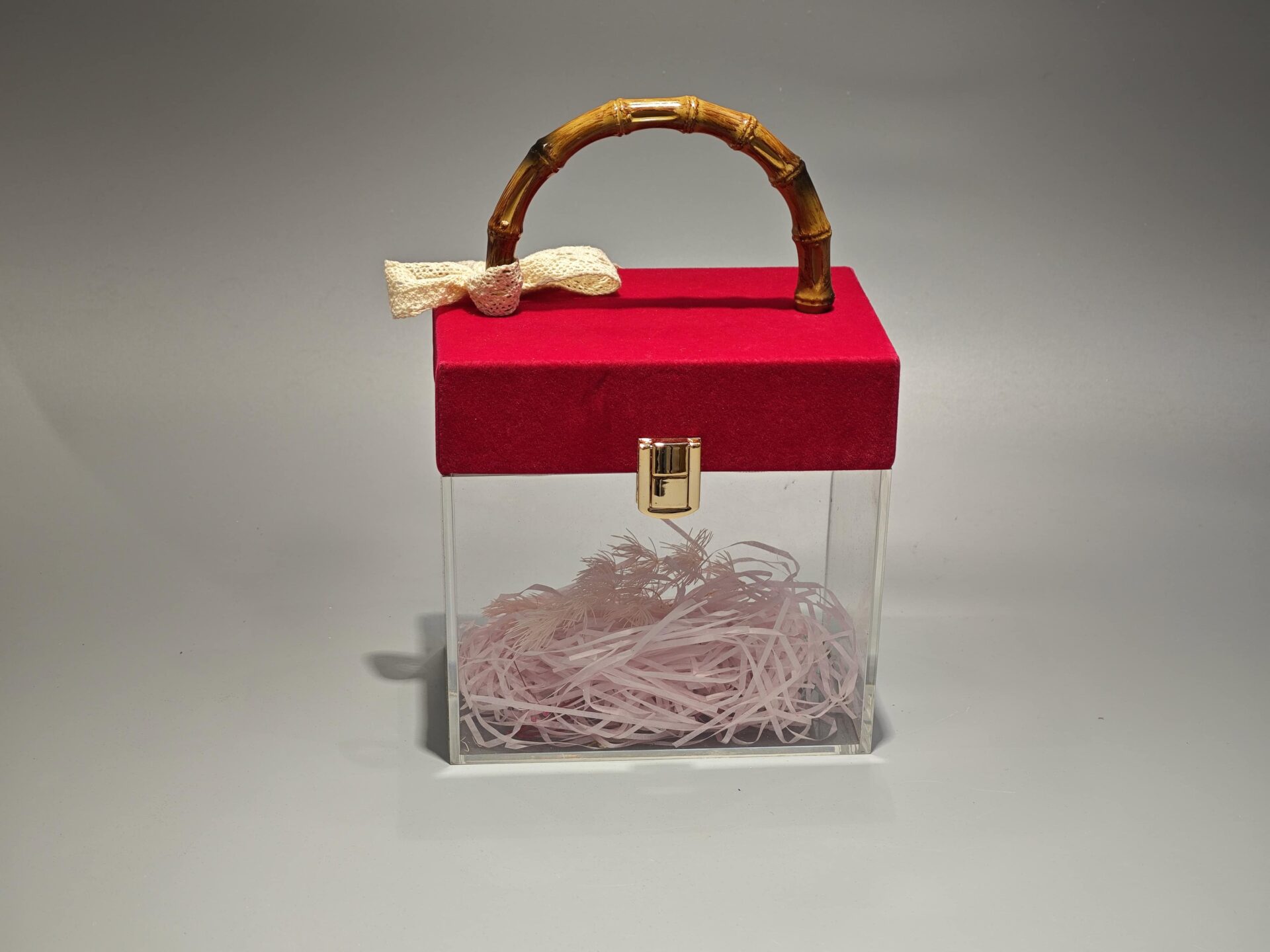A clear acrylic box featuring a flip-top lid covered in red velvet. It is adorned with a bamboo handle and decorated with a lace bow. The interior contains pink paper filler.