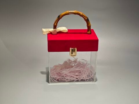 A clear acrylic box featuring a flip-top lid covered in red velvet. It is adorned with a bamboo handle and decorated with a lace bow. The interior contains pink paper filler.