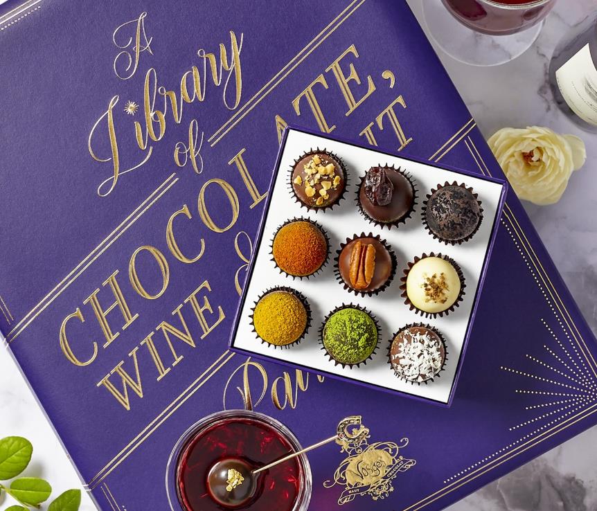 The image features a custom box of assorted chocolates placed on a purple book, accompanied by a glass of red wine and a white flower nearby.