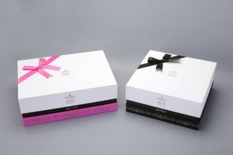 The image features two custom-designed packaging boxes adorned with decorative ribbons, one in pink and the other in black, set against a gray backdrop.
