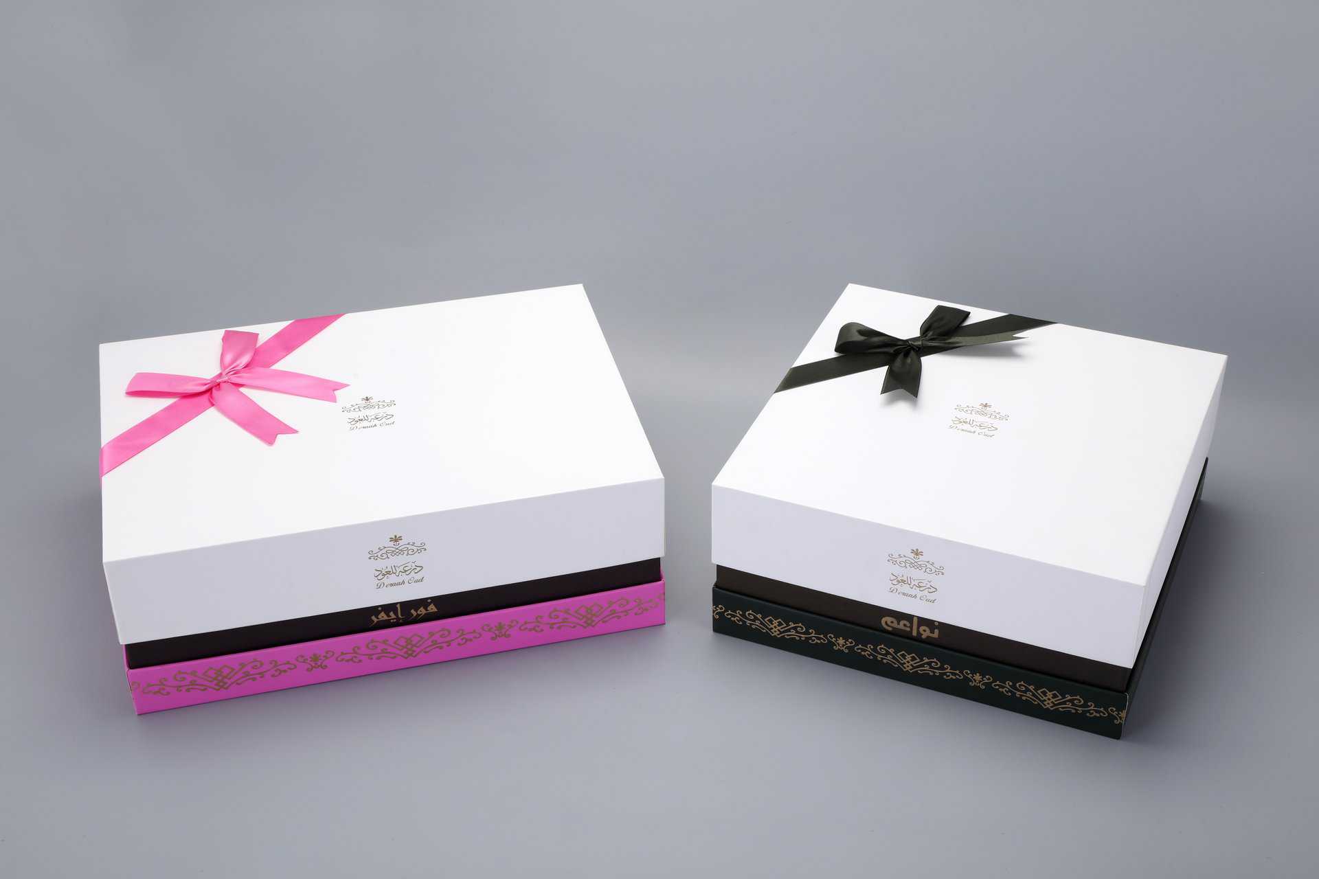 The image features two custom-designed packaging boxes adorned with decorative ribbons, one in pink and the other in black, set against a gray backdrop.
