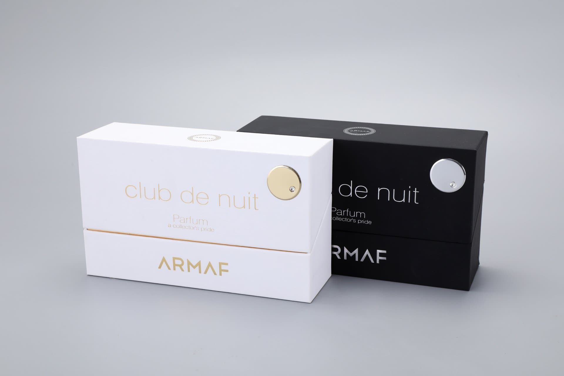 The image features two custom perfume packaging boxes by Armaf; one box is finished in black, while the other is in white. Both boxes are elegantly adorned with circular metallic embellishments, showcasing a sophisticated design.