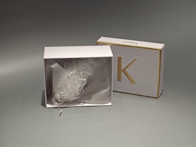 The image showcases a custom box in white and gold with a partially open lid, revealing white raffia inside.
