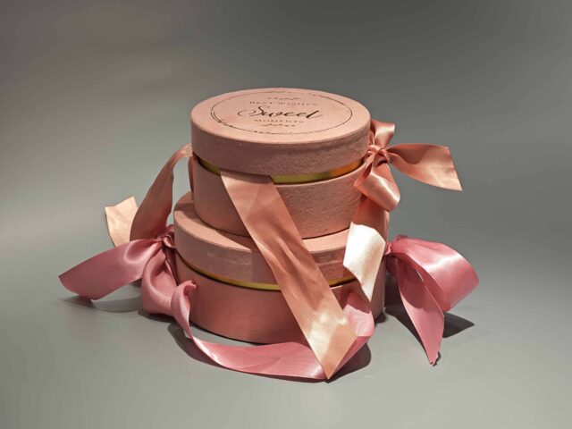 The image showcases a pair of custom round pink packaging boxes, designed with prominent pink satin ribbon bows, elegantly arranged on a gray surface.