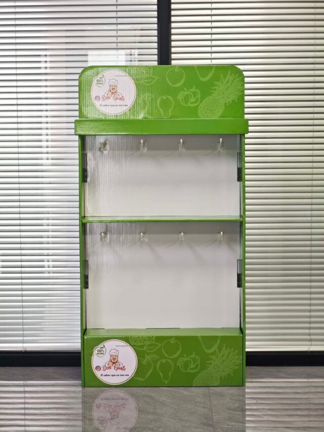The image shows a green, assembled display box with hanging slots positioned in a room with closed blinds in the background.