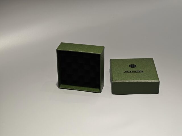 The product is a small green custom box designed with a lift-off lid, suitable for packaging needs. The box features minimalist branding on the front and is set against a plain background.