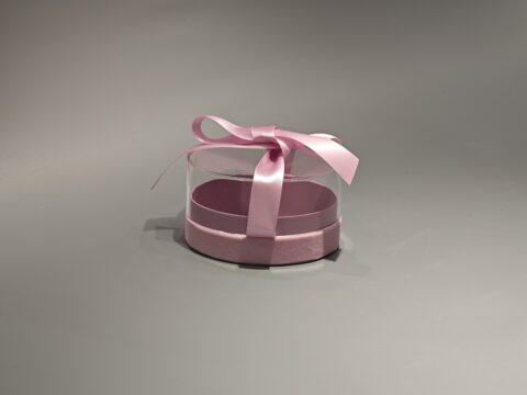 The product is a ellipse, pink custom box with a transparent lid, secured with a pink ribbon