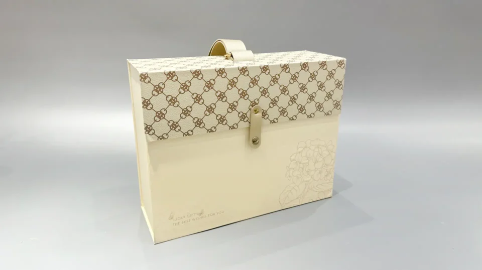 This beige packaging box showcases a patterned top panel and an elegant floral design on the bottom right corner, finished with a string and button closure.
