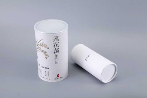 Two cylindrical tea canisters, one positioned upright featuring intricate floral designs and the other lying horizontally, displayed on a neutral gray background.