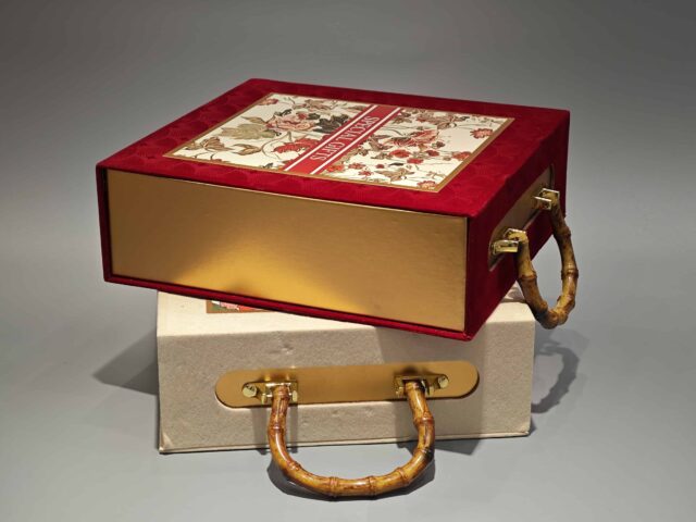 Two decorative packaging boxes feature unique bamboo handles. One box showcases a vibrant red color with an elegant floral design on its lid, while the other is a minimalist plain beige. Designed for easy portability and ideal for gift-giving, these boxes embody elegance in simplicity and are styled against a neutral gray background to highlight their refined appearance.