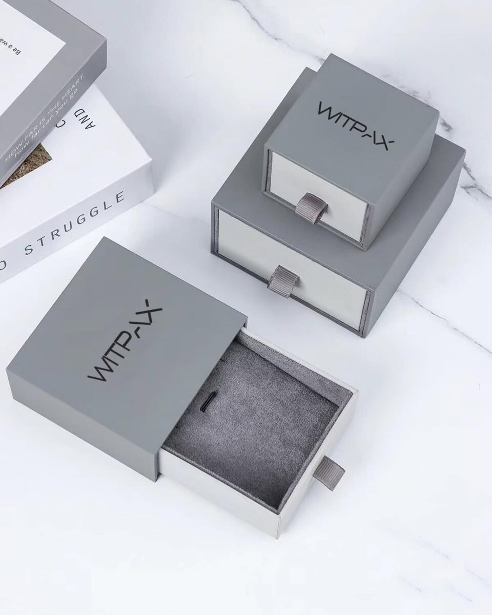 Two gray sliding drawer boxes are displayed, one open to reveal a soft interior. A glimpse of a book appears at the side. primary packaging example