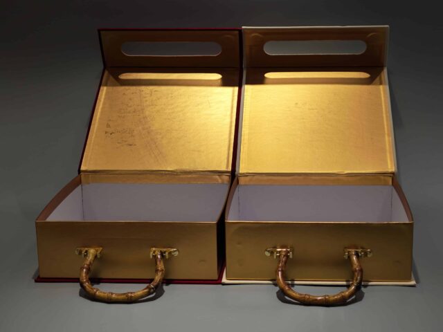 Two decorative packaging boxes feature unique bamboo handles. One box showcases a vibrant red color with an elegant floral design on its lid, while the other is a minimalist plain beige. Designed for easy portability and ideal for gift-giving, these boxes embody elegance in simplicity and are styled against a neutral gray background to highlight their refined appearance.