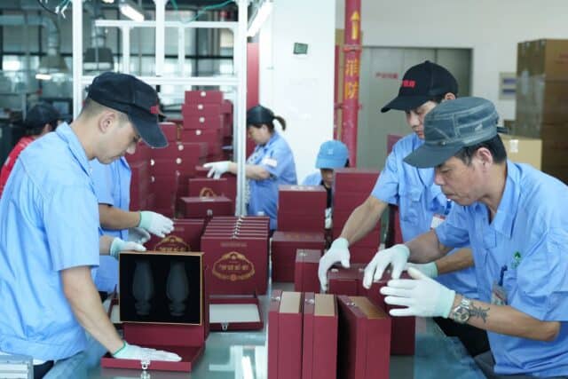 Workers in a factory assembling red boxes with velvet lining, wearing blue uniforms and gloves.