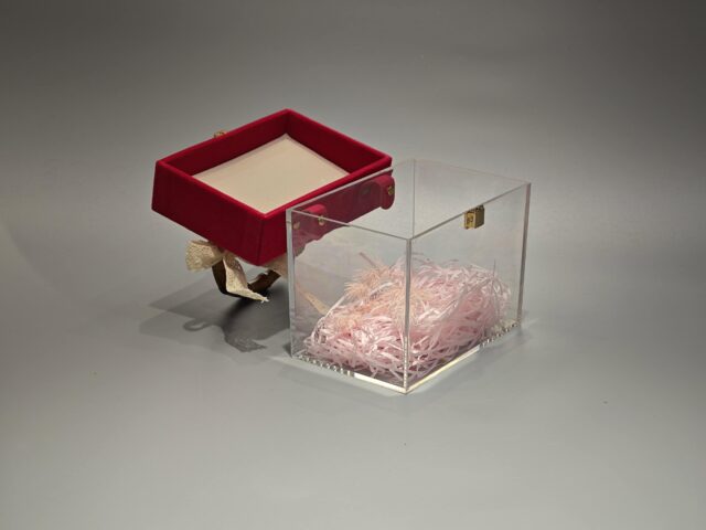 A clear acrylic box featuring a flip-top lid covered in red velvet. It is adorned with a bamboo handle and decorated with a lace bow. The interior contains pink paper filler.