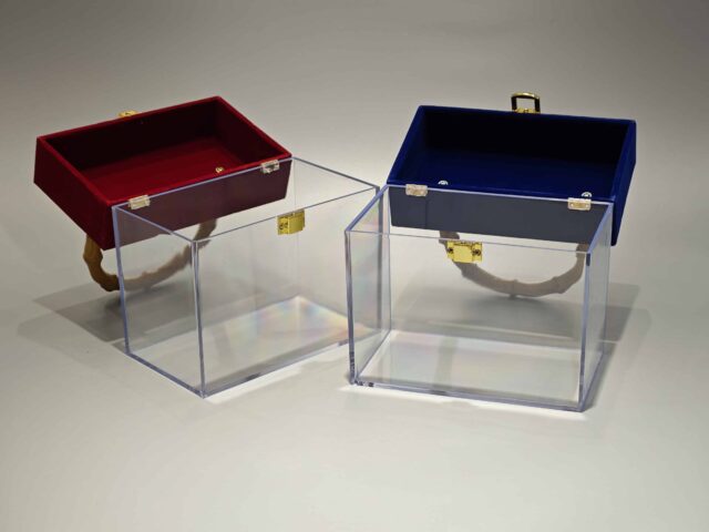 A clear acrylic box featuring a flip-top lid covered in red velvet. It is adorned with a bamboo handle and decorated with a lace bow. The interior contains pink paper filler.