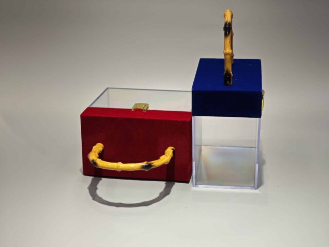 A clear acrylic box featuring a flip-top lid covered in red velvet. It is adorned with a bamboo handle and decorated with a lace bow. The interior contains pink paper filler.