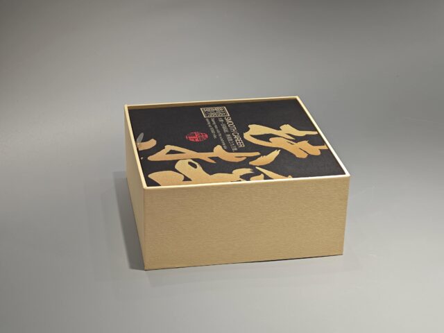 A packaging box with a handle, adorned with black Asian calligraphy and an intricate patterned design.