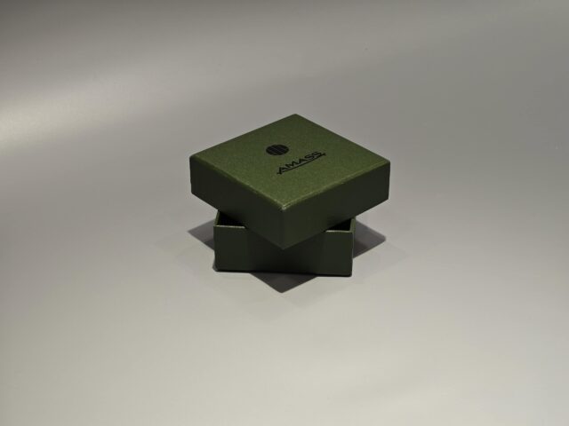 The product is a small green custom box designed with a lift-off lid, suitable for packaging needs. The box features minimalist branding on the front and is set against a plain background.