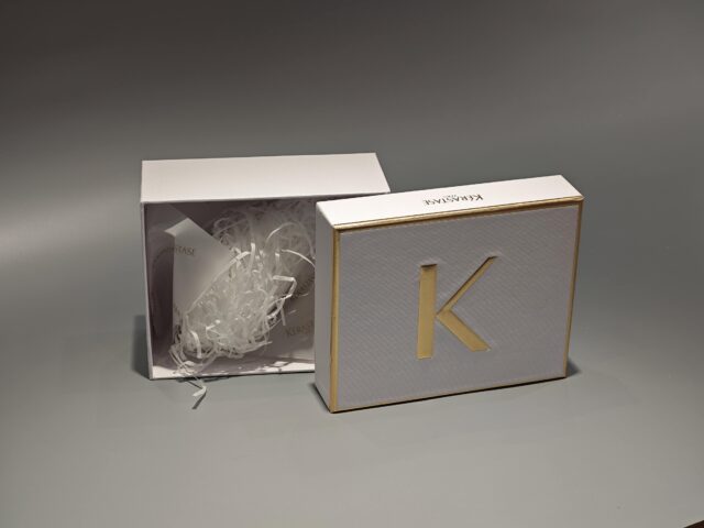 The image showcases a custom box in white and gold with a partially open lid, revealing white raffia inside.