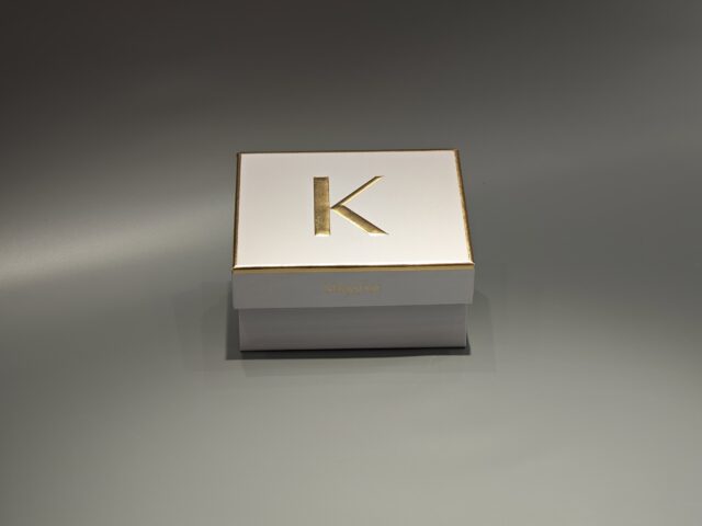 The image showcases a custom box in white and gold with a partially open lid, revealing white raffia inside.