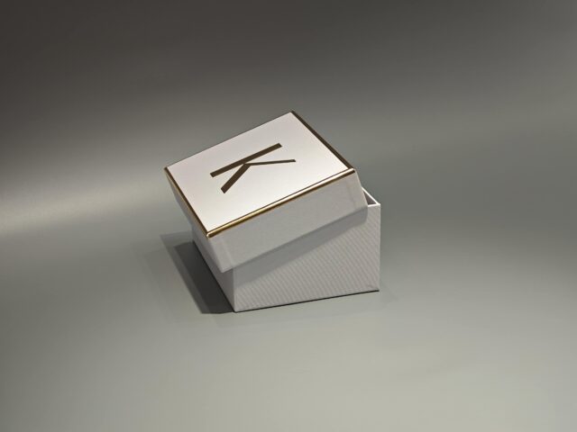 The image showcases a custom box in white and gold with a partially open lid, revealing white raffia inside.