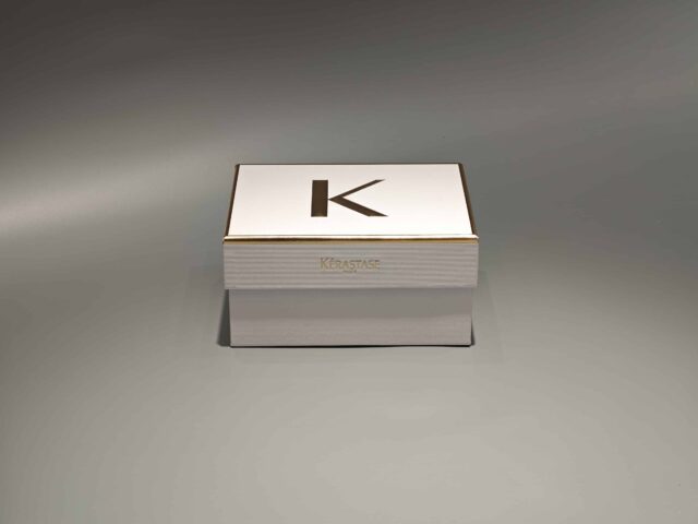 The image showcases a custom box in white and gold with a partially open lid, revealing white raffia inside.