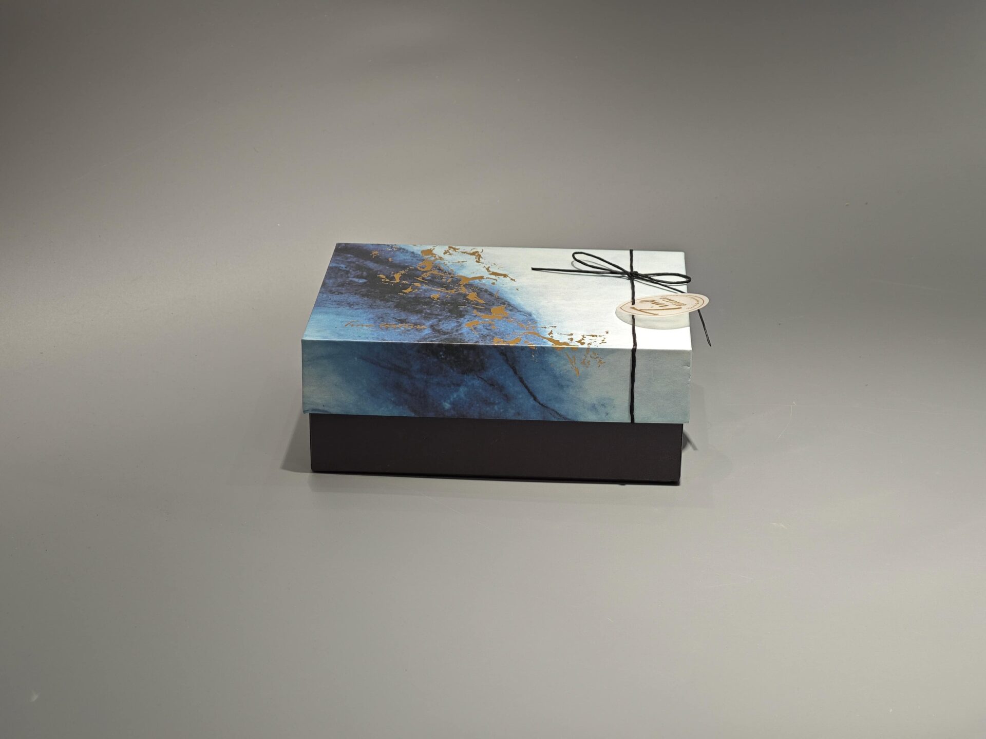 This custom ribbon box showcases a striking blue and gold abstract pattern. Designed with a lift-off lid, it is elegantly finished with a black cord and includes a round tag for added personalization.