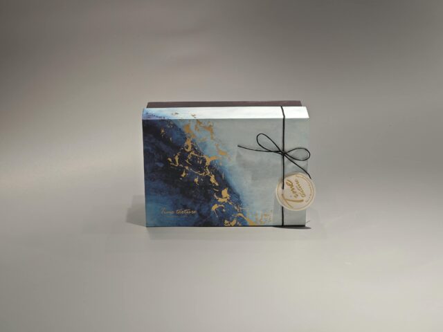 This custom ribbon box showcases a striking blue and gold abstract pattern. Designed with a lift-off lid, it is elegantly finished with a black cord and includes a round tag for added personalization.