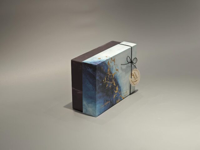 This custom ribbon box showcases a striking blue and gold abstract pattern. Designed with a lift-off lid, it is elegantly finished with a black cord and includes a round tag for added personalization.