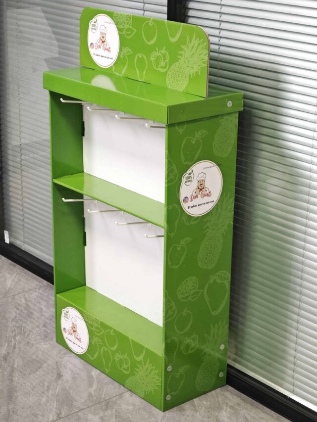 The image shows a green, assembled display box with hanging slots positioned in a room with closed blinds in the background.