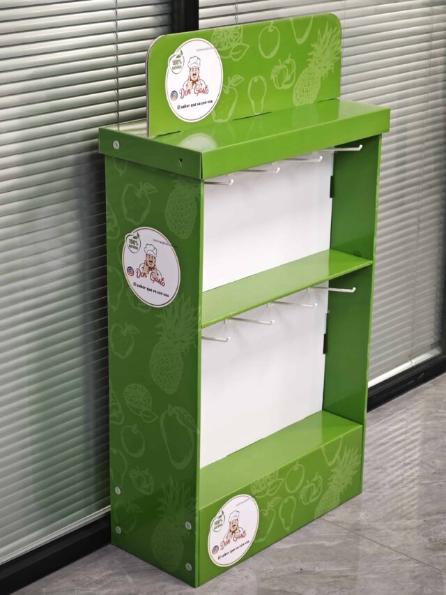 The image shows a green, assembled display box with hanging slots positioned in a room with closed blinds in the background.