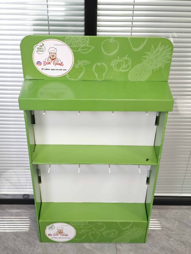 The image shows a green, assembled display box with hanging slots positioned in a room with closed blinds in the background.