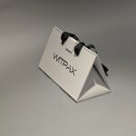 The custom collapsible packaging box, crafted from white cardboard and featuring sleek black handles, sits elegantly on a light gray surface, exemplifying sophisticated design.