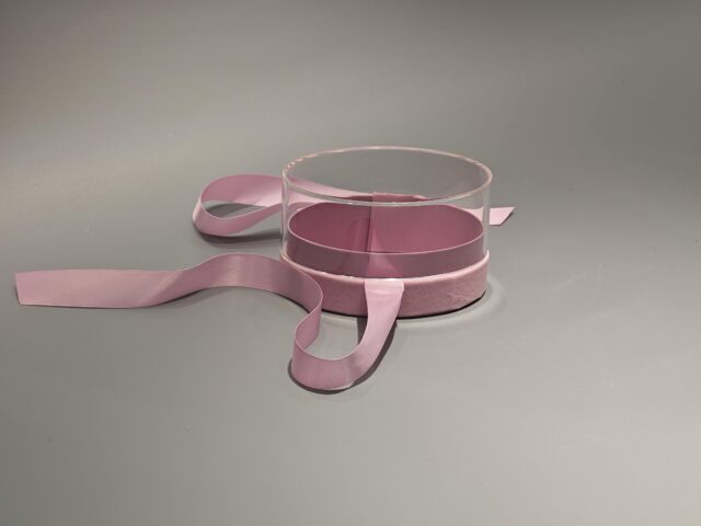 The product is a ellipse, pink custom box with a transparent lid, secured with a pink ribbon