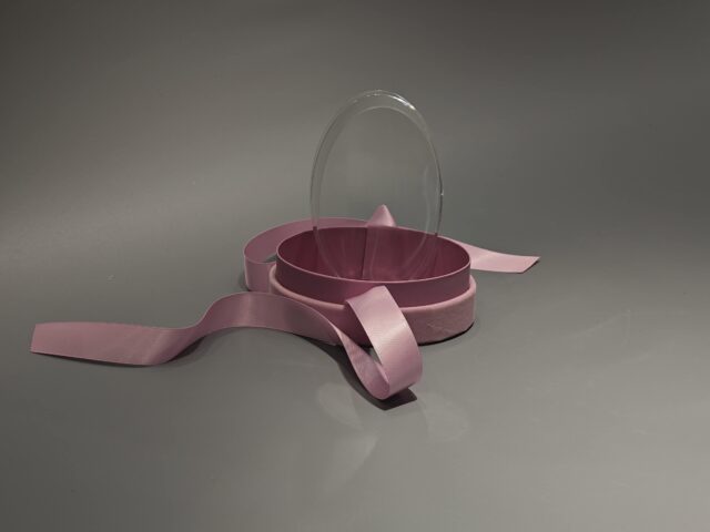 The product is a ellipse, pink custom box with a transparent lid, secured with a pink ribbon