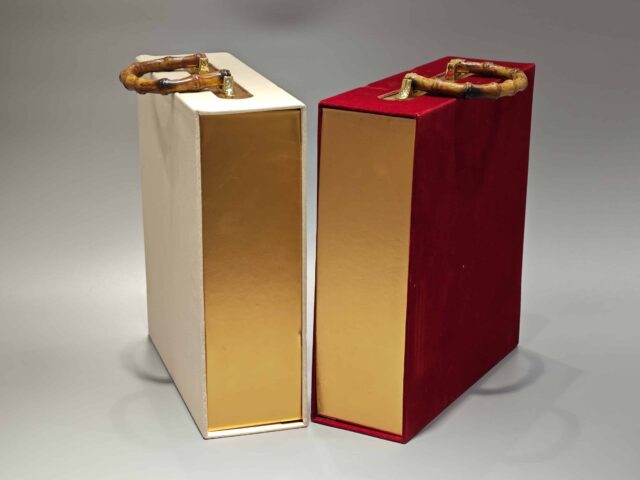 Two decorative packaging boxes feature unique bamboo handles. One box showcases a vibrant red color with an elegant floral design on its lid, while the other is a minimalist plain beige. Designed for easy portability and ideal for gift-giving, these boxes embody elegance in simplicity and are styled against a neutral gray background to highlight their refined appearance.